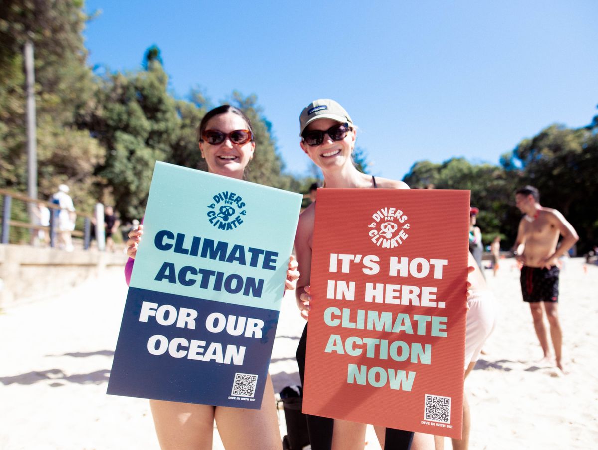 Two Divers for Climate supporters standing on a beach holding campaign signs that read Climate action for our ocean and It's hot in here. Climate action now