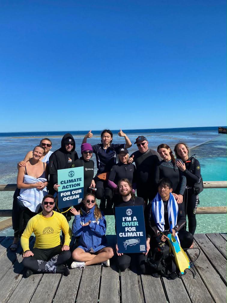A group of divers in wetsuits and rashguards pose on a wooden deck above turquoise reef water holding a Climate action for our ocean sign