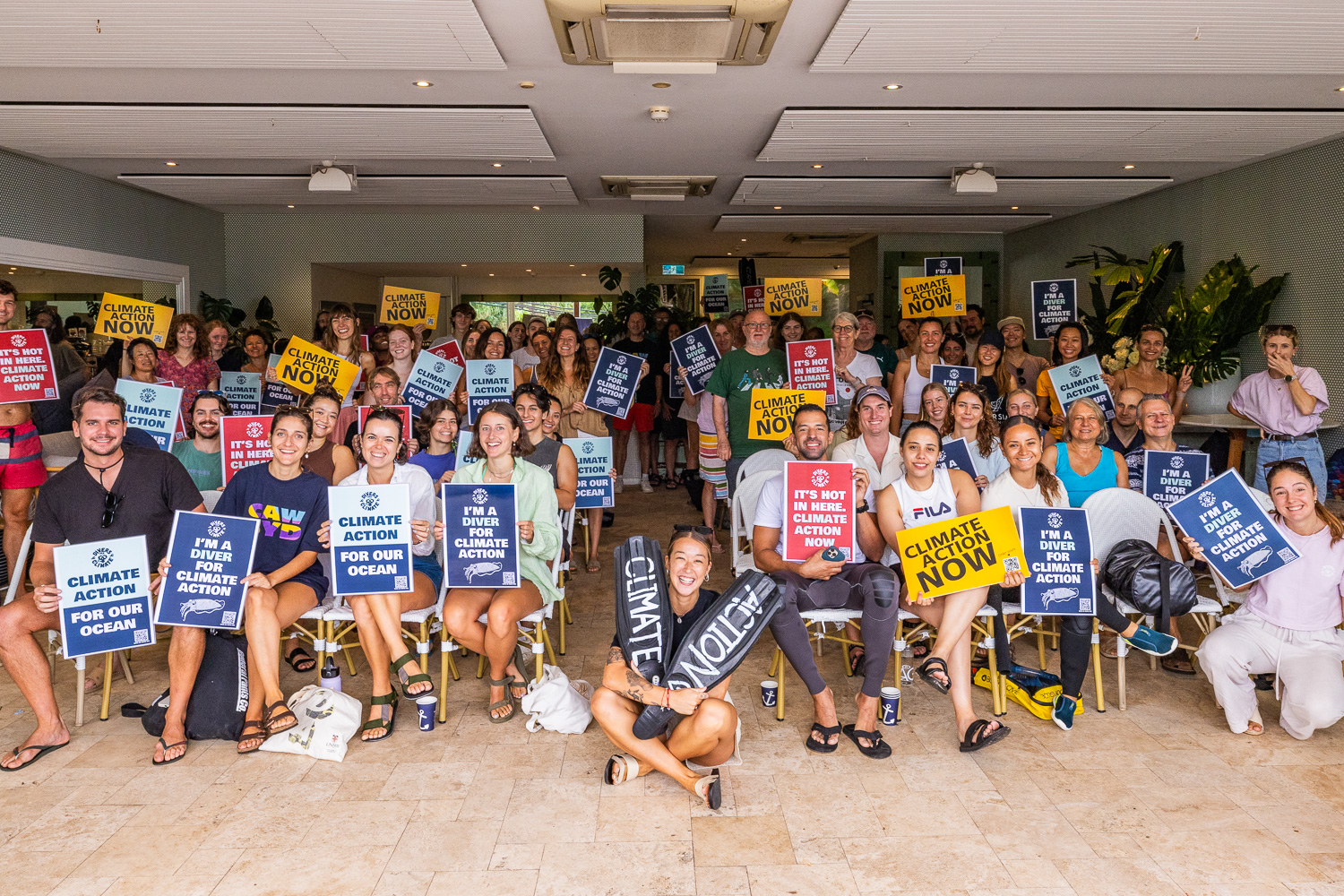 A large group of divers and climate advocates posing indoors with colourful signs reading Climate Action Now, I'm a Diver for Climate and Climate Action for Our Ocean