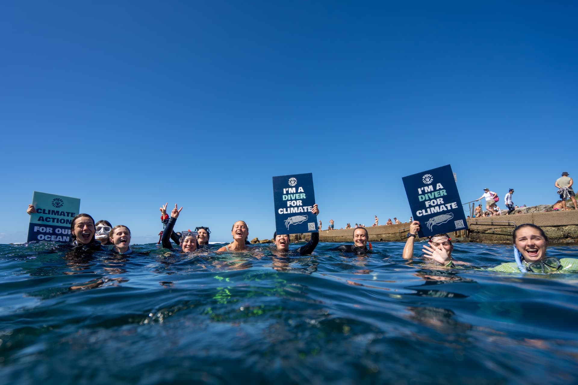 A group of snorkelers floating in the ocean smiling and holding up signs reading I'm a Diver for Climate and Climate Action for Our Ocean under a clear blue sky