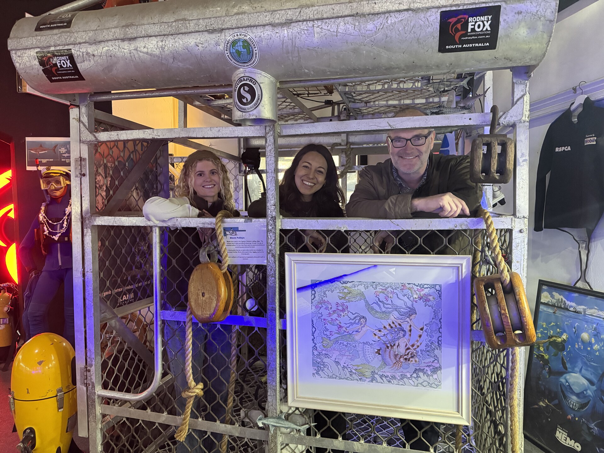 Three people smile from inside a metal shark diving cage at a South Australian museum exhibit, with a vintage diving suit and wooden pulleys nearby